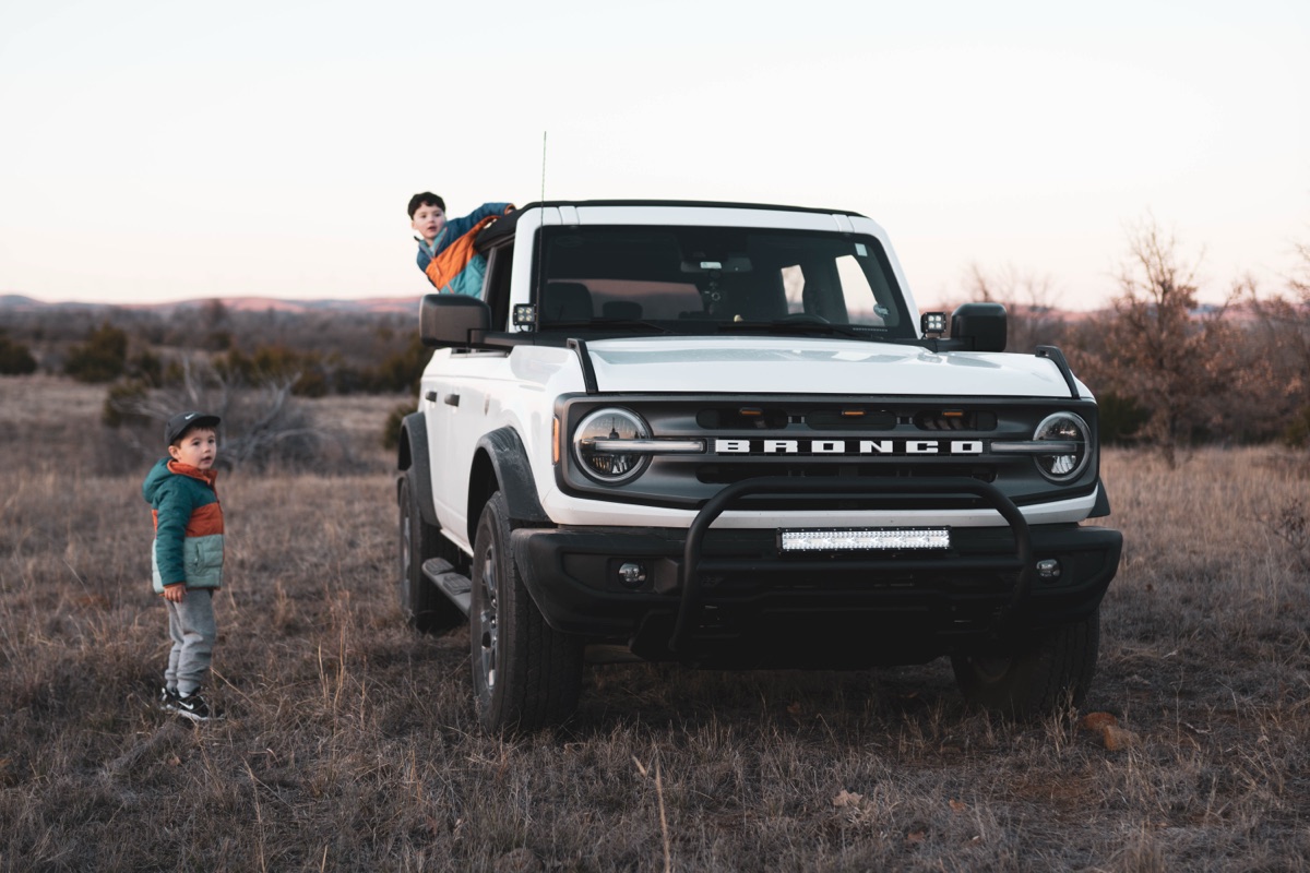Two kids and a white Ford Bronco in a pasture at dusk